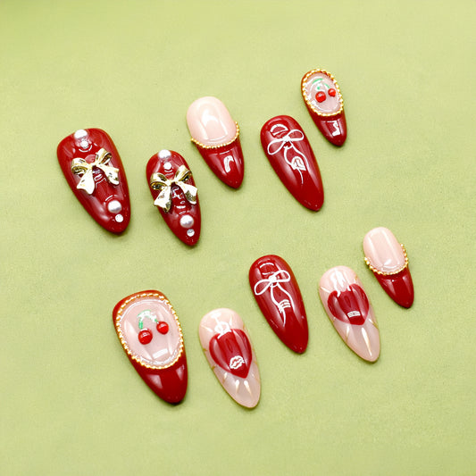 Cherry Kiss & Crimson Bows press-on nails with red and nude tones, 3D cherry and gold bow charms, heart and ribbon accents, almond shape.