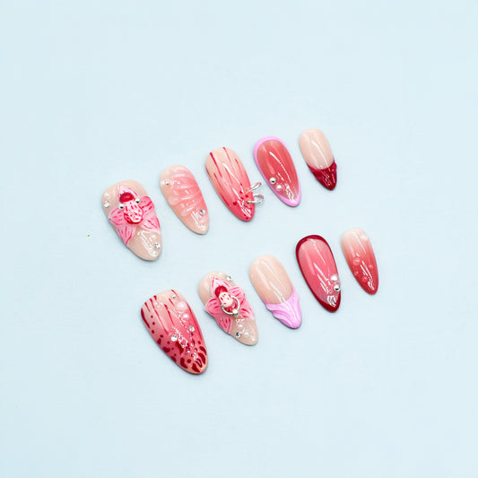 Medium Almond press-on nails in crimson and pink ombré with 3D orchid flowers, hand-painted streaks, and rhinestone accents.