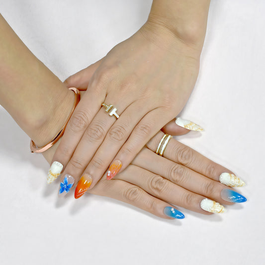 Handmade press-on nails in long almond shape featuring tropical flower designs, ombré orange and blue gradient, sunbeam linework, and pearl accents – Sunset Bloom set.