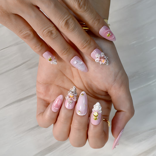 Medium almond press-on nails in soft pink with 3D floral appliqués, white botanical detailing, and gold seashell accents – Blush Bloom nail set.