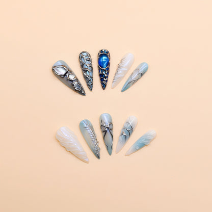 Long stiletto press-on nails in icy blue and silver with chrome textures, gemstone accents, and futuristic 3D design.