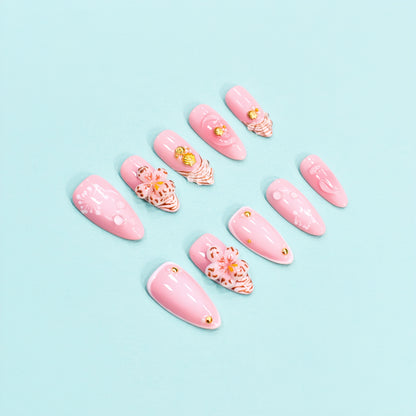 Medium almond press-on nails in soft pink with 3D floral appliqués, white botanical detailing, and gold seashell accents – Blush Bloom nail set.