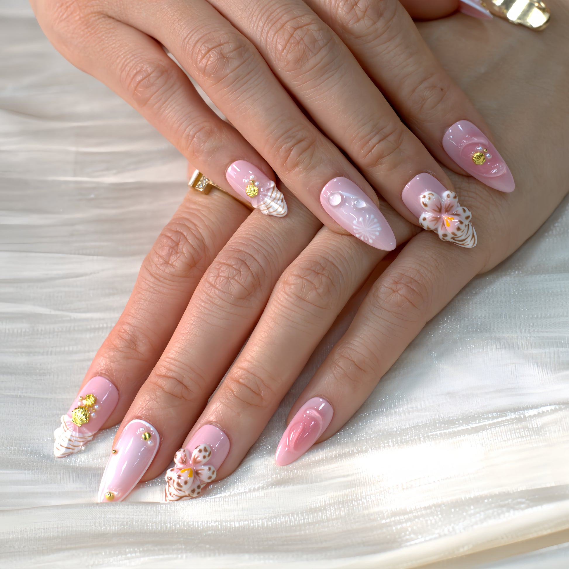 Medium almond press-on nails in soft pink with 3D floral appliqués, white botanical detailing, and gold seashell accents – Blush Bloom nail set.