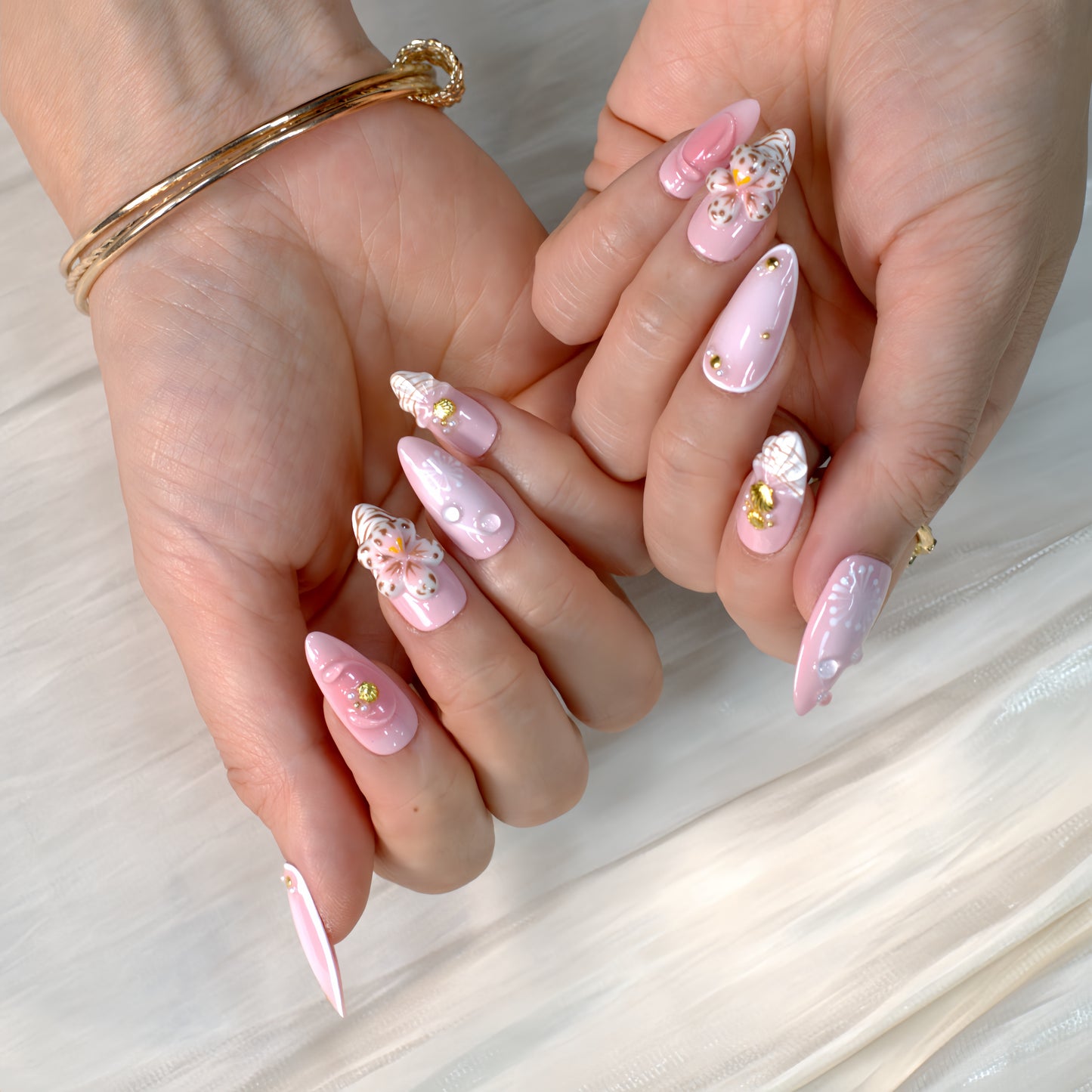 Medium almond press-on nails in soft pink with 3D floral appliqués, white botanical detailing, and gold seashell accents – Blush Bloom nail set.