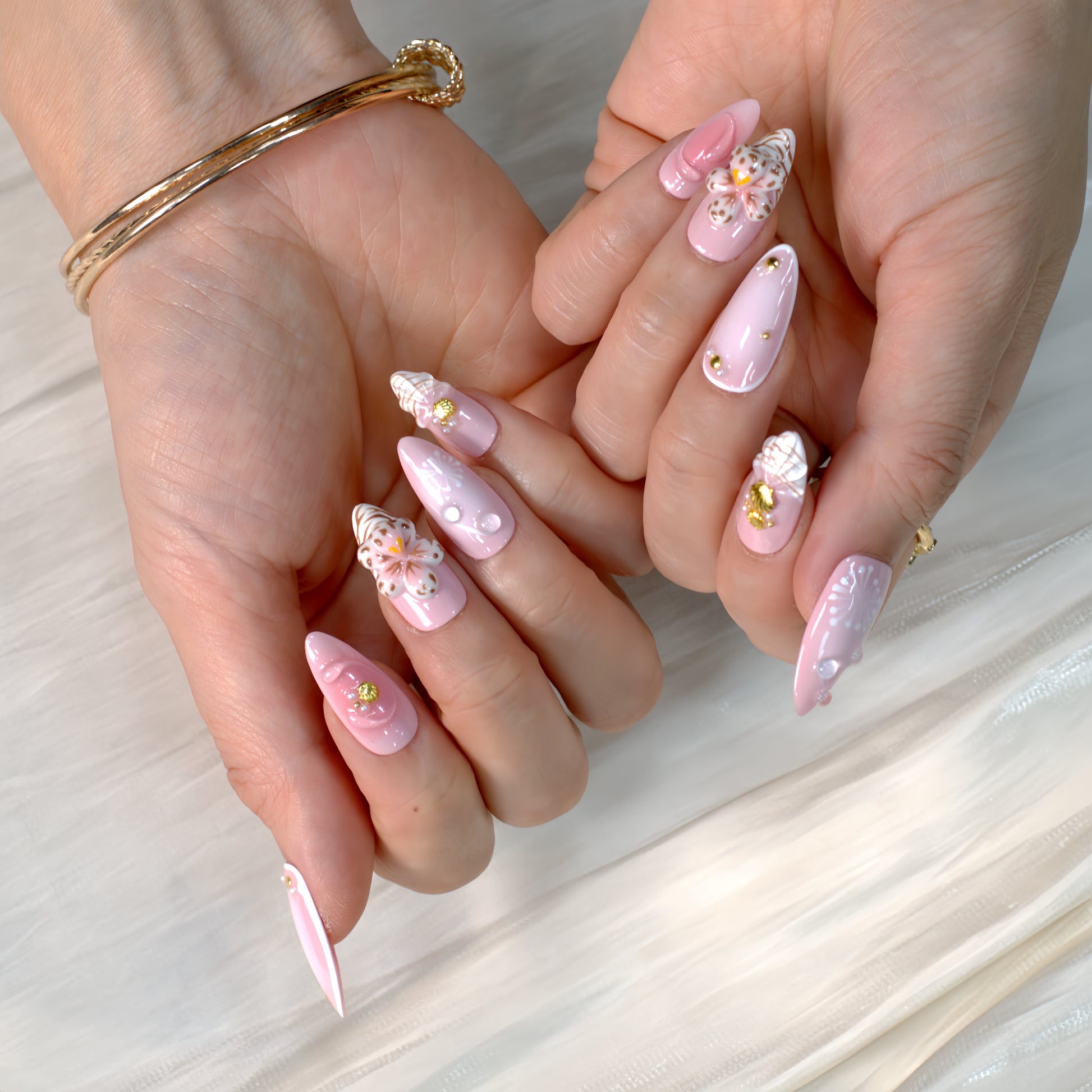 Medium almond press-on nails in soft pink with 3D floral appliqués, white botanical detailing, and gold seashell accents – Blush Bloom nail set.