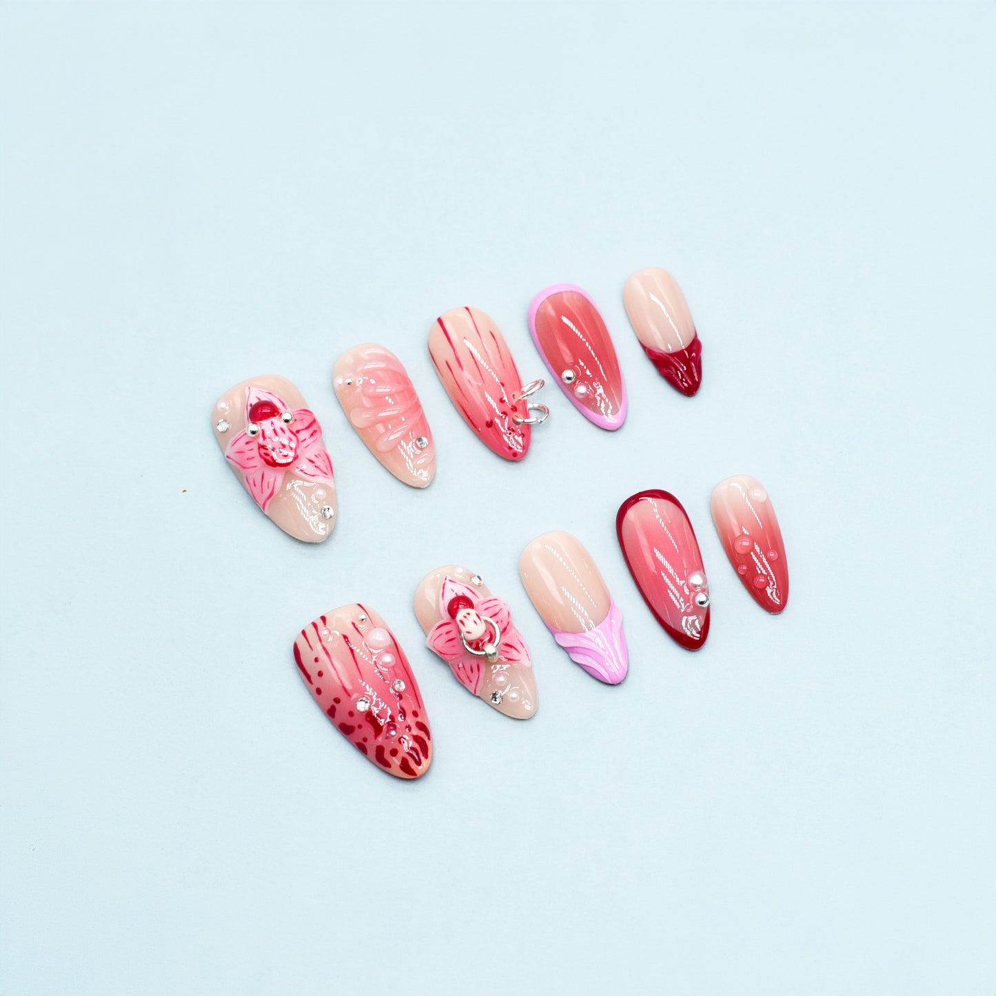 Medium Almond press-on nails in crimson and pink ombré with 3D orchid flowers, hand-painted streaks, and rhinestone accents.