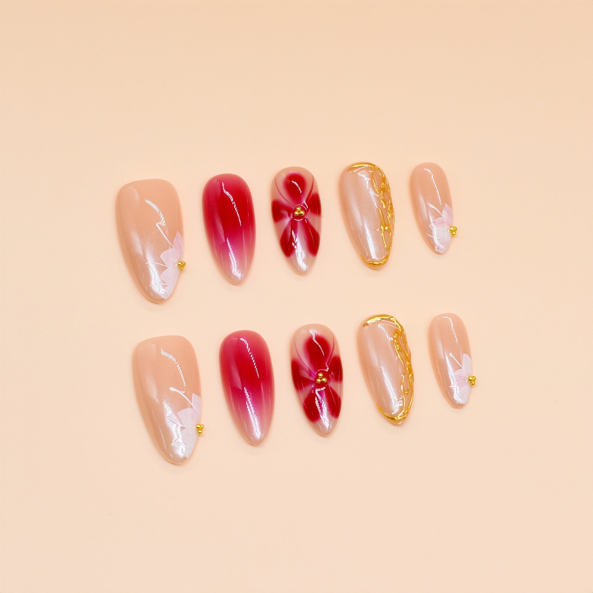 Medium almond press-on nails in nude and crimson ombré with white florals, gold beads, and sculpted gold frame.