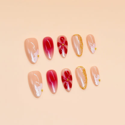 Medium almond press-on nails in nude and crimson ombré with white florals, gold beads, and sculpted gold frame.