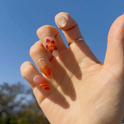 Golden Ember Bloom press-on nails in amber-orange with gold contour lines and red floral 3D accents, captured under blue sky lighting.