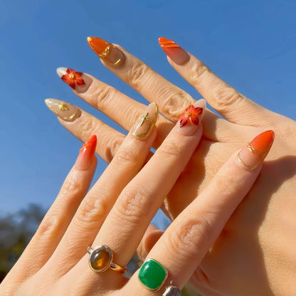 Golden Ember Bloom press-on nails in amber-orange with gold contour lines and red floral 3D accents, captured under blue sky lighting.