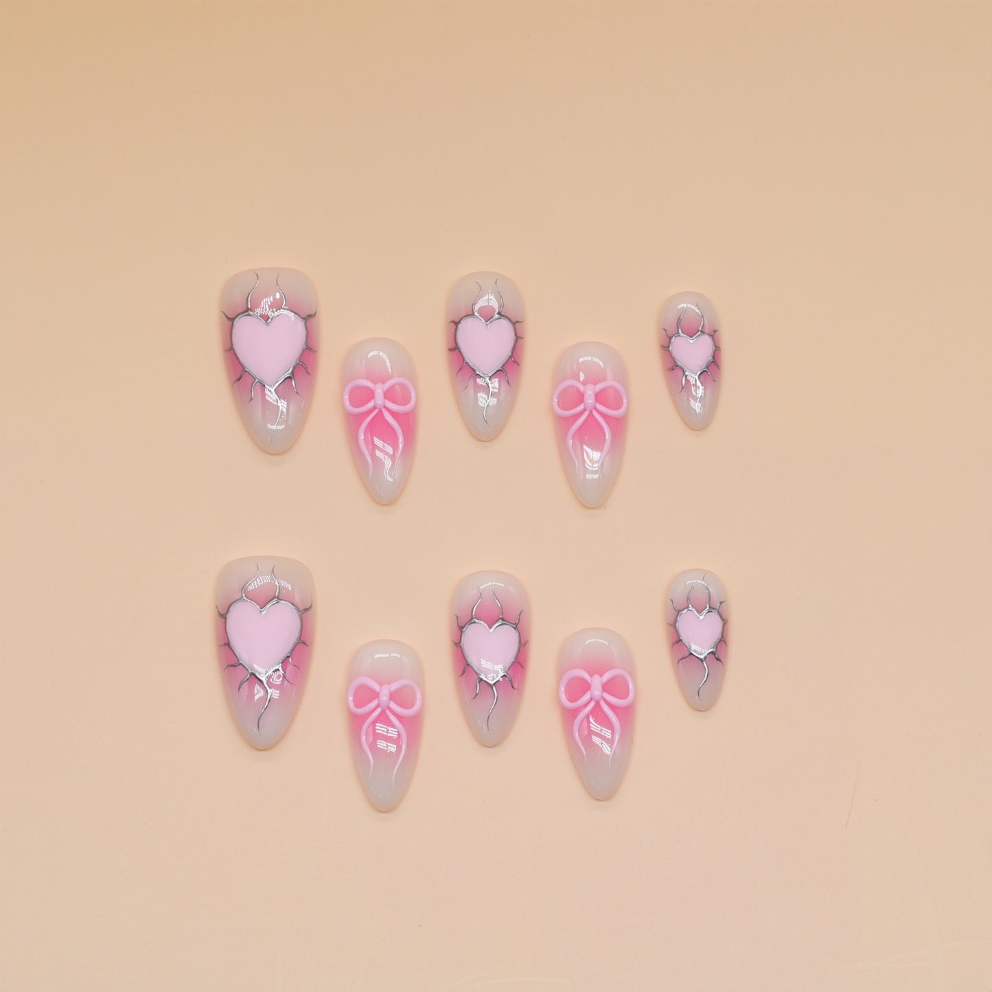 Medium almond press-on nails with nude-to-pink ombré, 3D pink hearts, bows, and crack detailing.