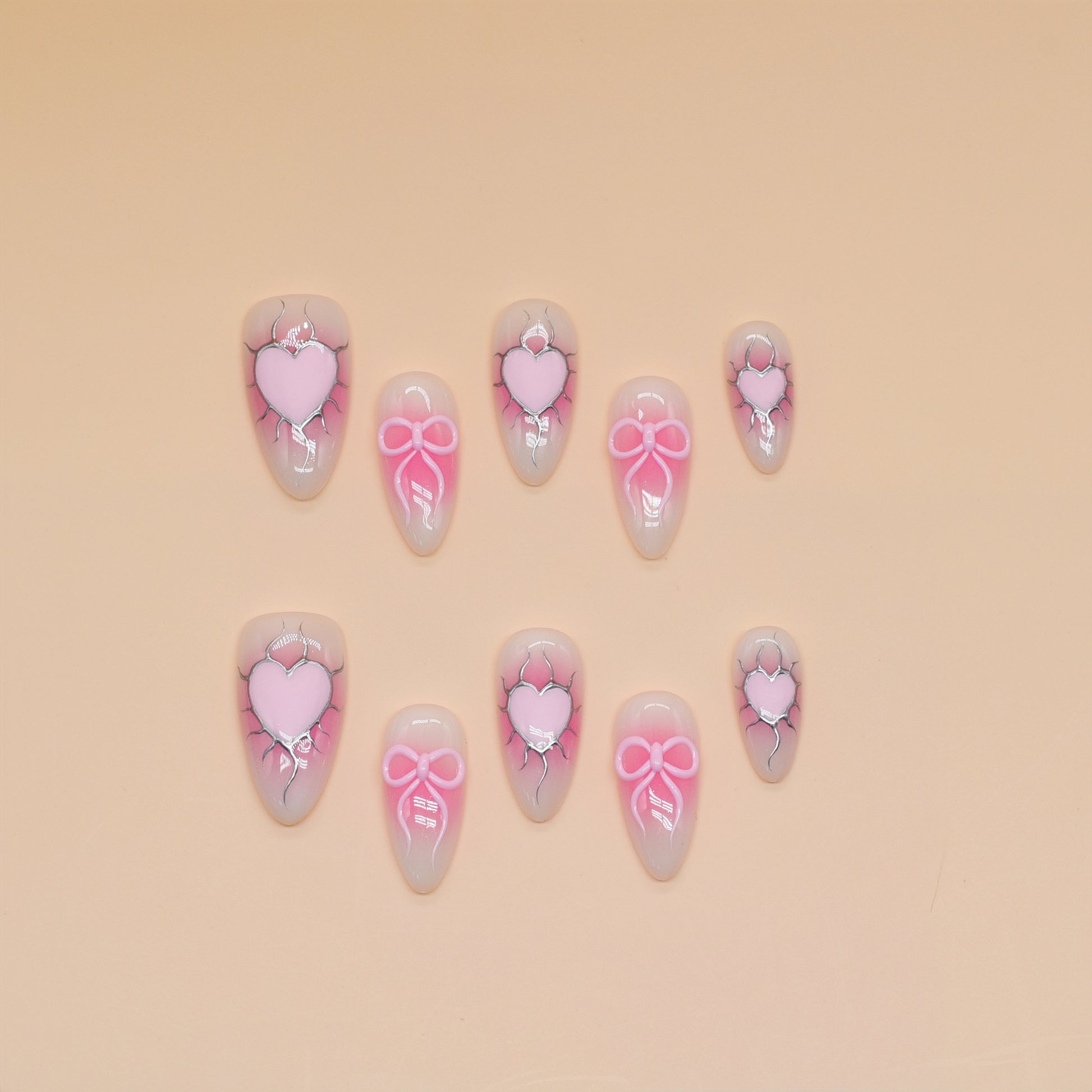 Medium almond press-on nails with nude-to-pink ombré, 3D pink hearts, bows, and crack detailing.