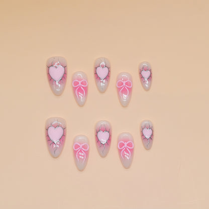 Medium almond press-on nails with nude-to-pink ombré, 3D pink hearts, bows, and crack detailing.
