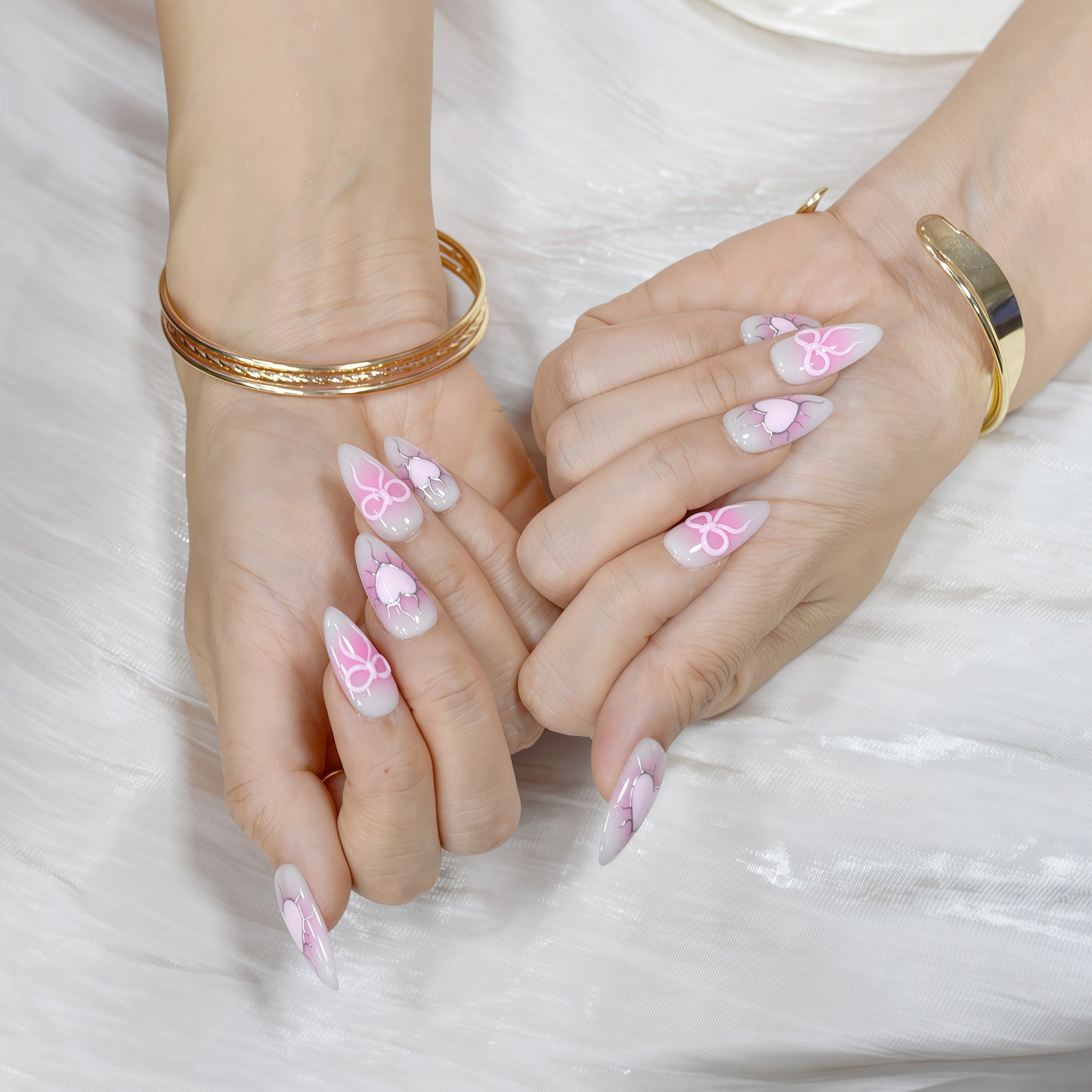 Medium almond press-on nails with nude-to-pink ombré, 3D pink hearts, bows, and crack detailing.