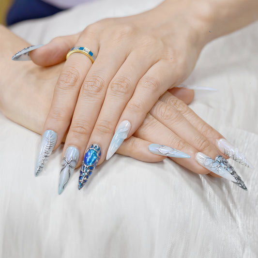 Long stiletto press-on nails in icy blue and silver with chrome textures, gemstone accents, and futuristic 3D design.