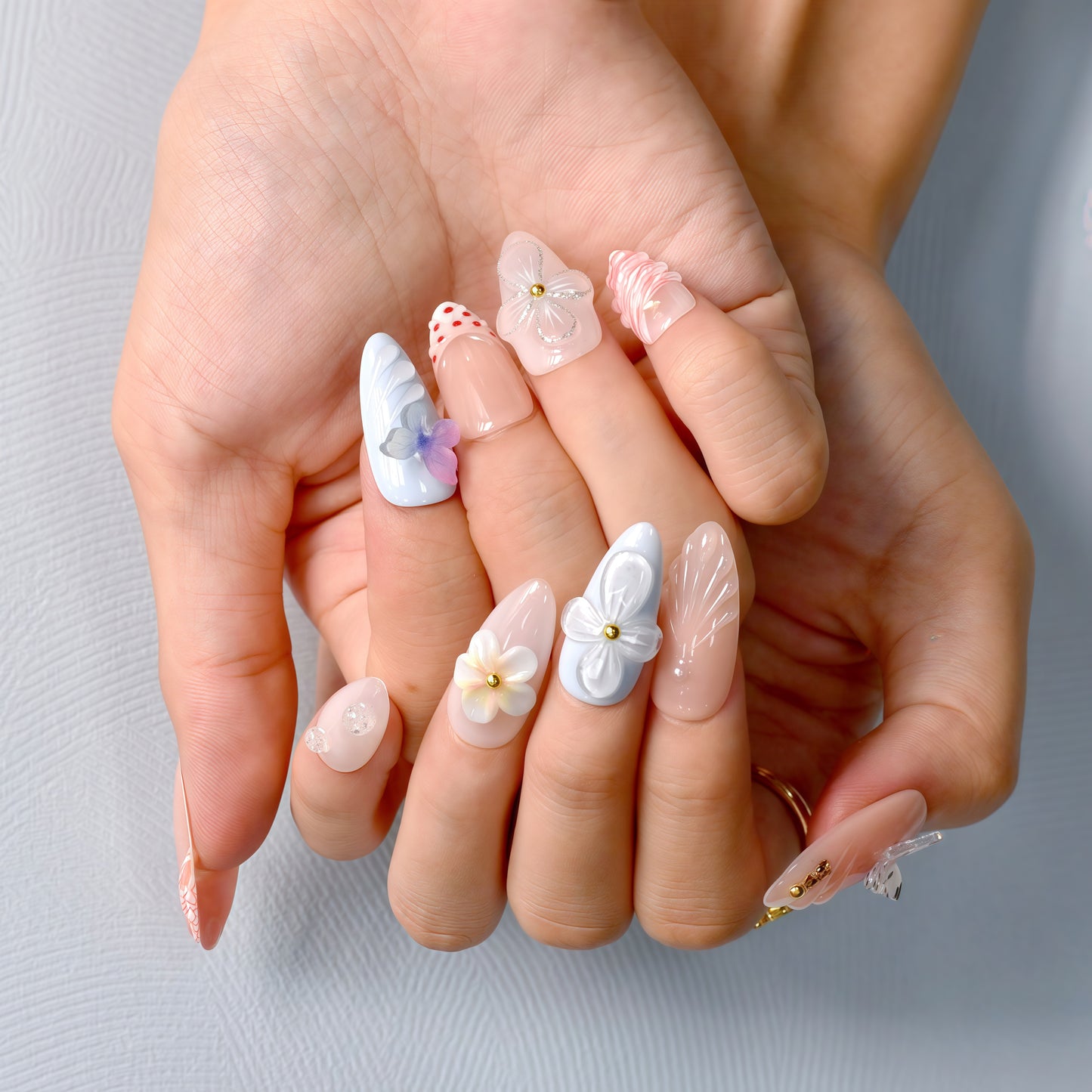 press-on nails in pastel blue, coral, nude, and white with 3D flowers, butterflies, bows, and glitter accents.