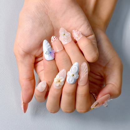 press-on nails in pastel blue, coral, nude, and white with 3D flowers, butterflies, bows, and glitter accents.