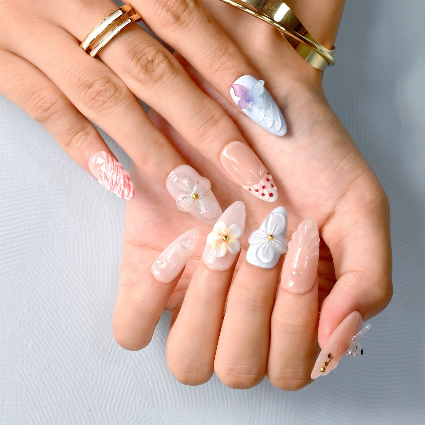 press-on nails in pastel blue, coral, nude, and white with 3D flowers, butterflies, bows, and glitter accents.