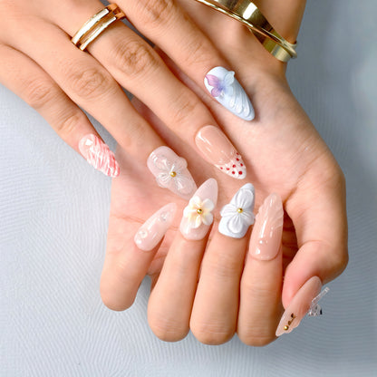 press-on nails in pastel blue, coral, nude, and white with 3D flowers, butterflies, bows, and glitter accents.