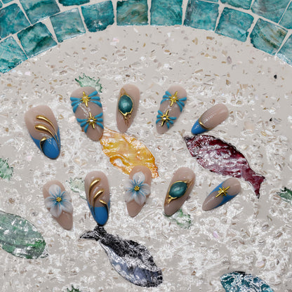 Handmade ocean blue press on nails with gold starfish charms, floral details and gemstone accents on nude base.