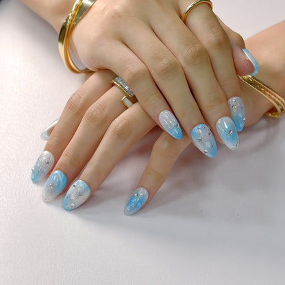Blue gradient press-on nails with white ombre tips, starburst designs, and rhinestone details displayed on a soft textured background