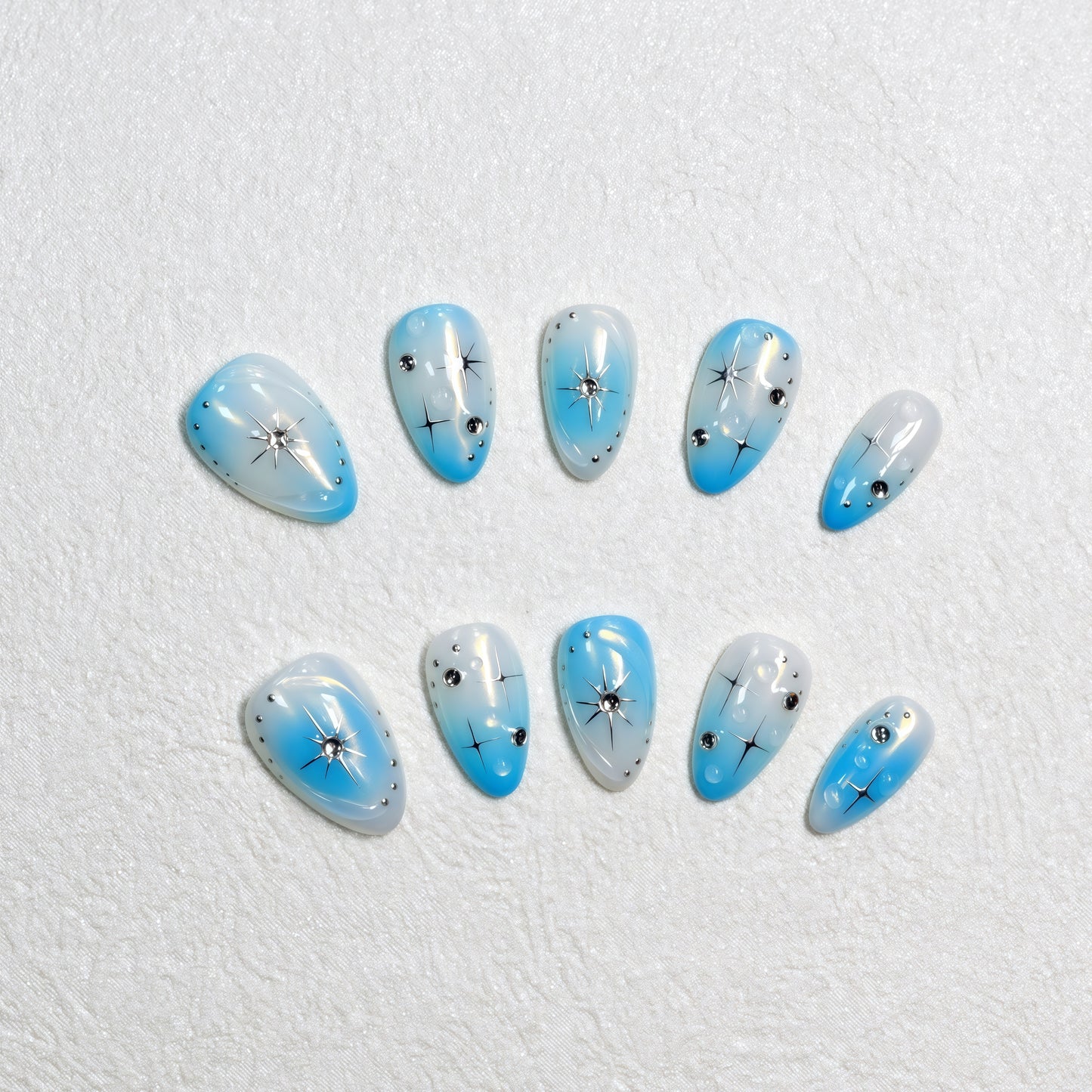 Blue gradient press-on nails with white ombre tips, starburst designs, and rhinestone details displayed on a soft textured background