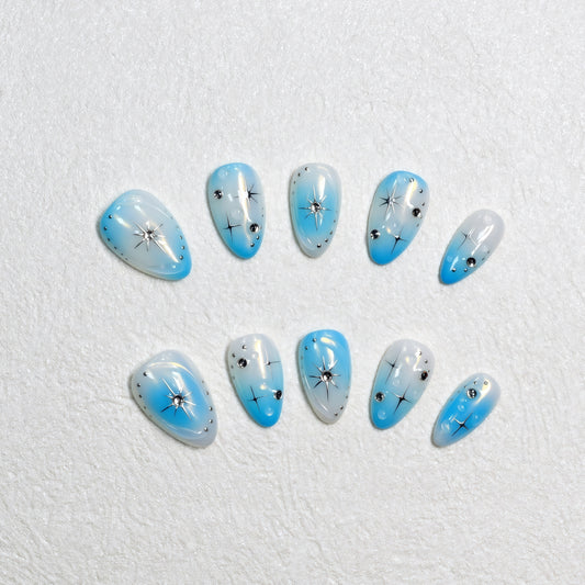 Blue gradient press-on nails with white ombre tips, starburst designs, and rhinestone details displayed on a soft textured background