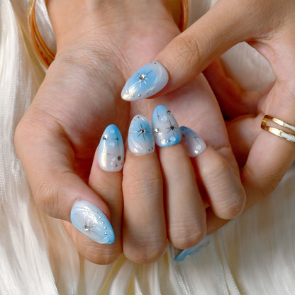 Blue gradient press-on nails with white ombre tips, starburst designs, and rhinestone details displayed on a soft textured background