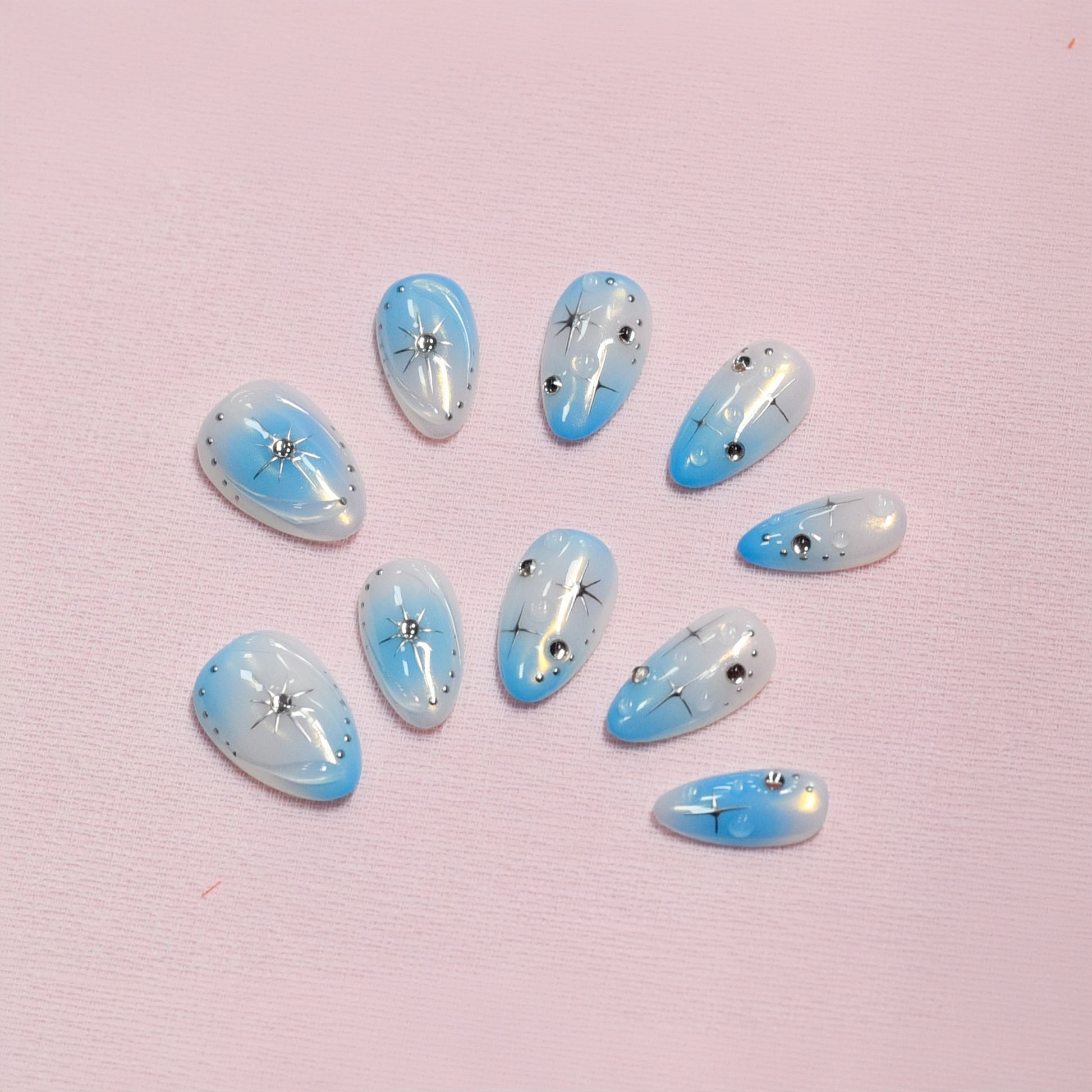 Blue gradient press-on nails with white ombre tips, starburst designs, and rhinestone details displayed on a soft textured background
