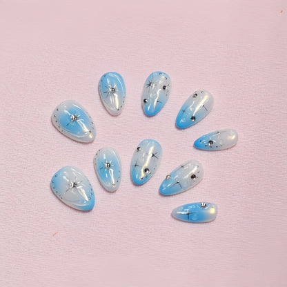 Blue gradient press-on nails with white ombre tips, starburst designs, and rhinestone details displayed on a soft textured background
