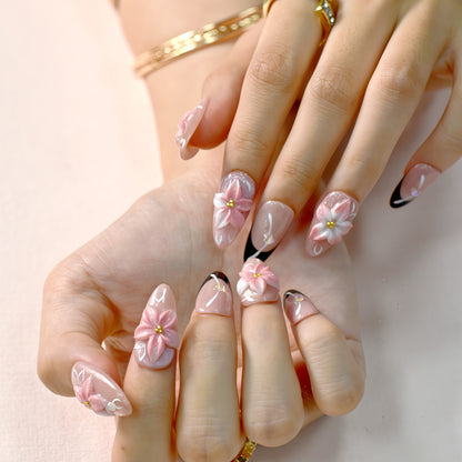 Luxury pink floral press-on nails with 3D flower design, pearl embellishments, and black French tip accents displayed on beige textured background