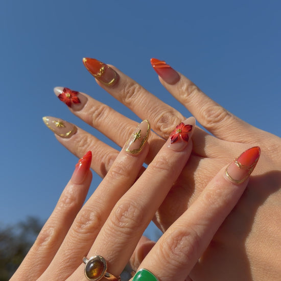 Golden Ember Bloom press-on nails in amber-orange with gold contour lines and red floral 3D accents, captured under blue sky lighting.