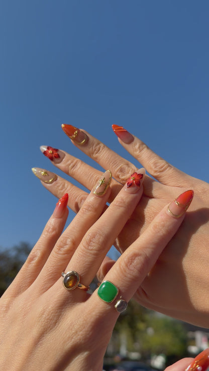 Golden Ember Bloom press-on nails in amber-orange with gold contour lines and red floral 3D accents, captured under blue sky lighting.